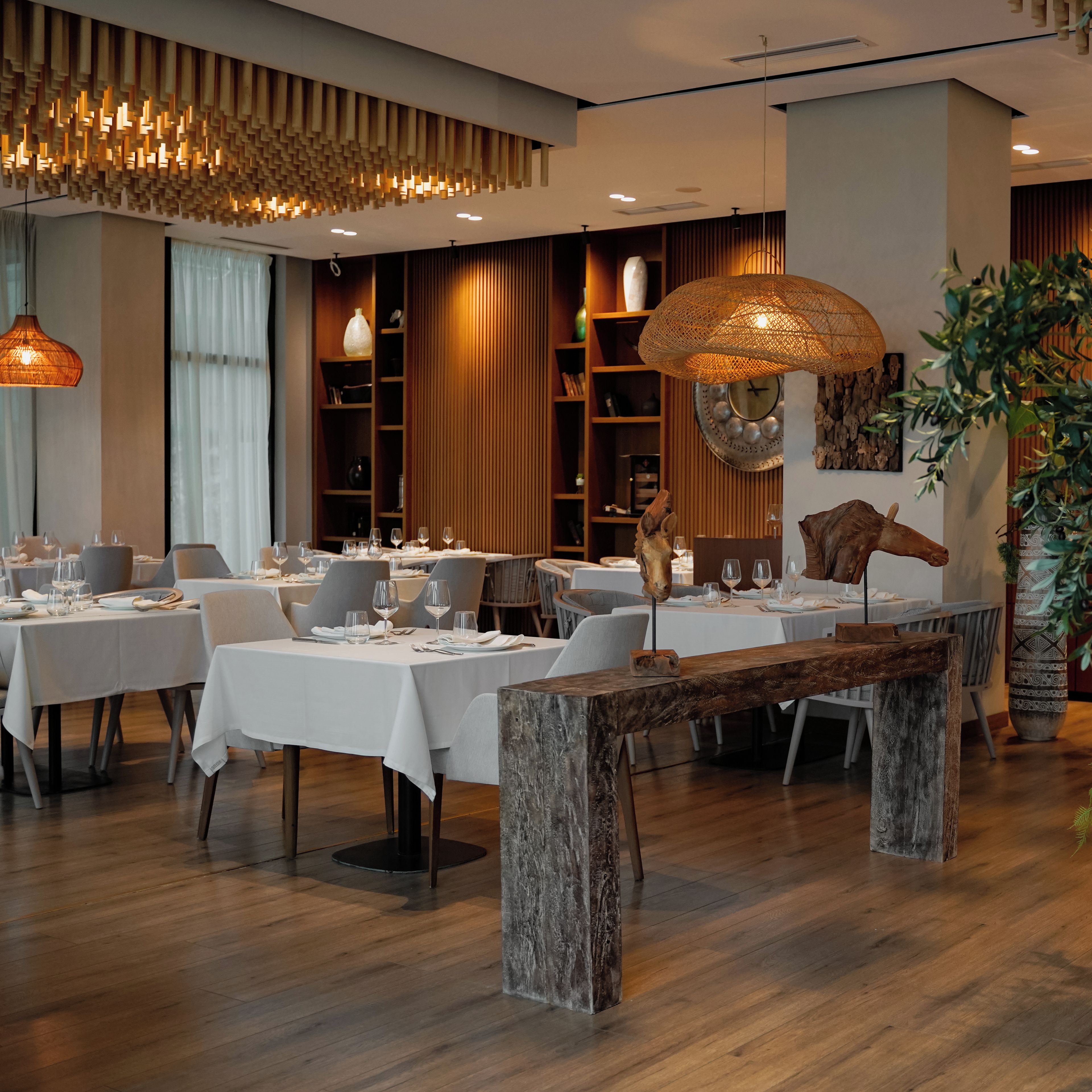 A modern dining area with wooden tables, white tablecloths, and a variety of decorative items including a large woven basket and a wooden sculpture.
