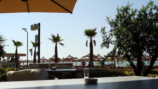 An outdoor dining area with tables and chairs, palm trees, and umbrellas. - Supreme Hotel (Golem)