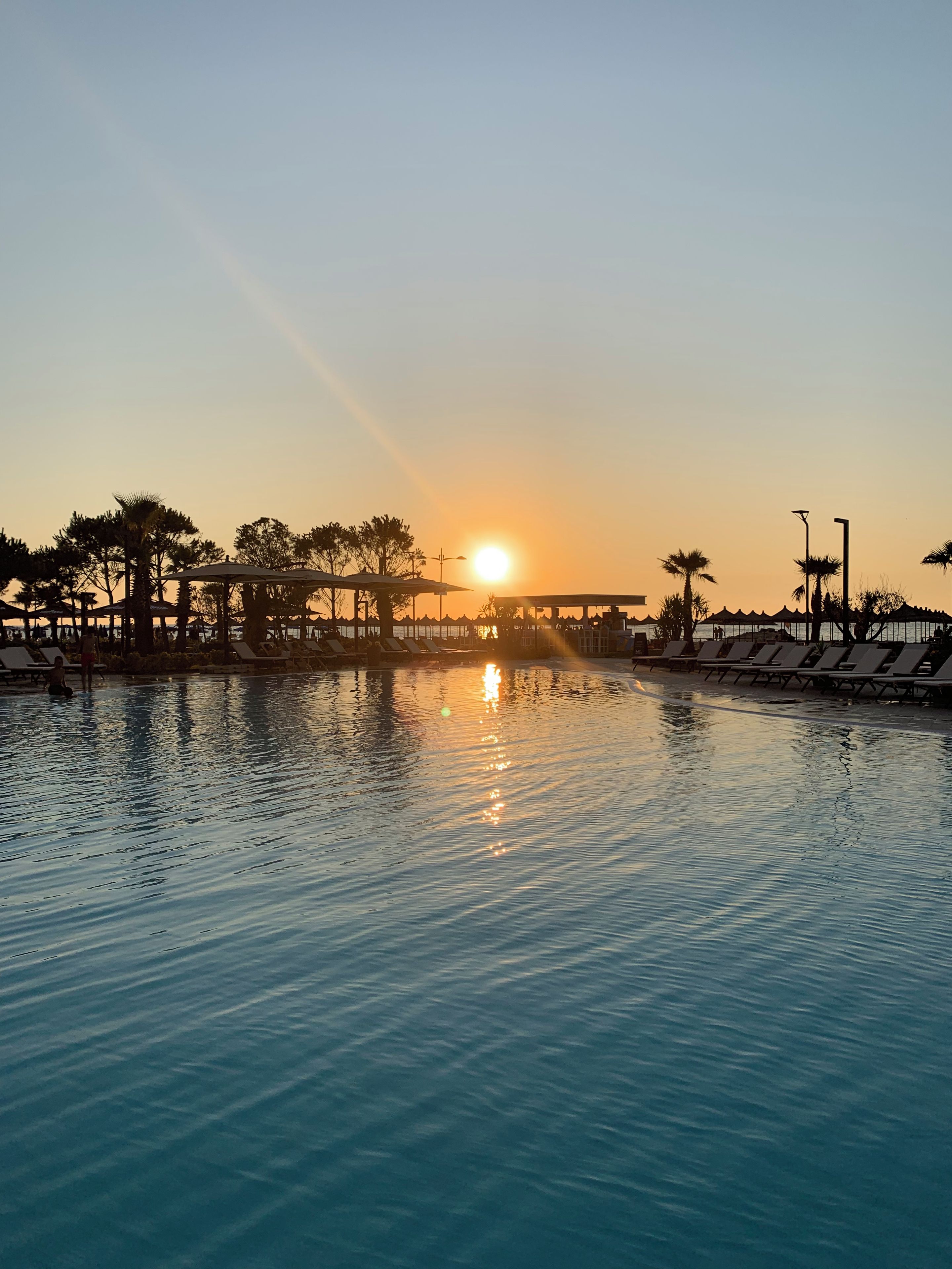 A swimming pool with lounge chairs and umbrellas, surrounded by trees and a sunset in the background.