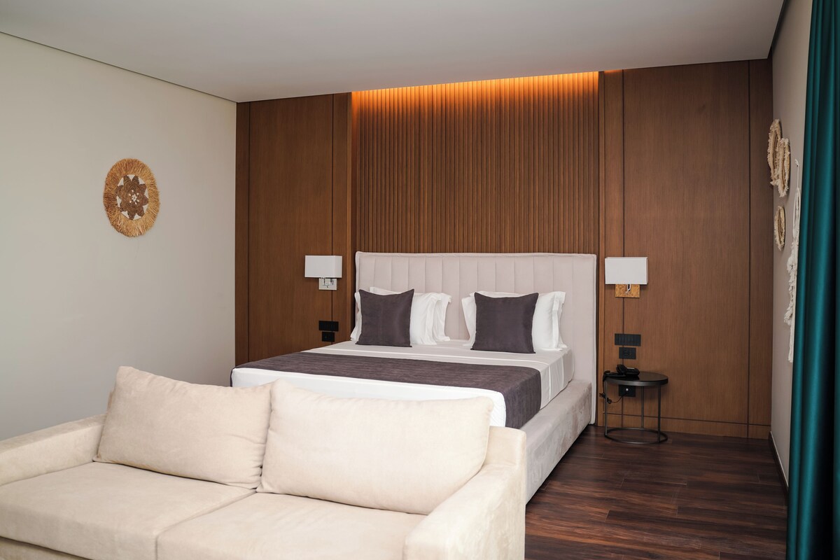 A modern hotel room with a large bed, two bedside tables with lamps, a sofa, and a decorative wall piece.