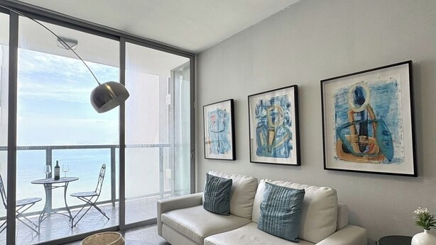 Un salon moderne avec un canapé blanc, un fauteuil bleu et une table ronde agrémentée d’objets décoratifs. Trois œuvres d’art encadrées ornent le mur, et une grande fenêtre offre une vue sur la mer.