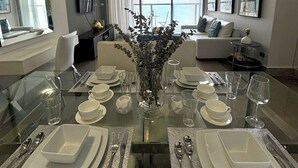 Un espace repas moderne avec une table en verre pouvant accueillir six personnes, un vase décoré de verdure et une vue sur la mer grâce à de grandes fenêtres.