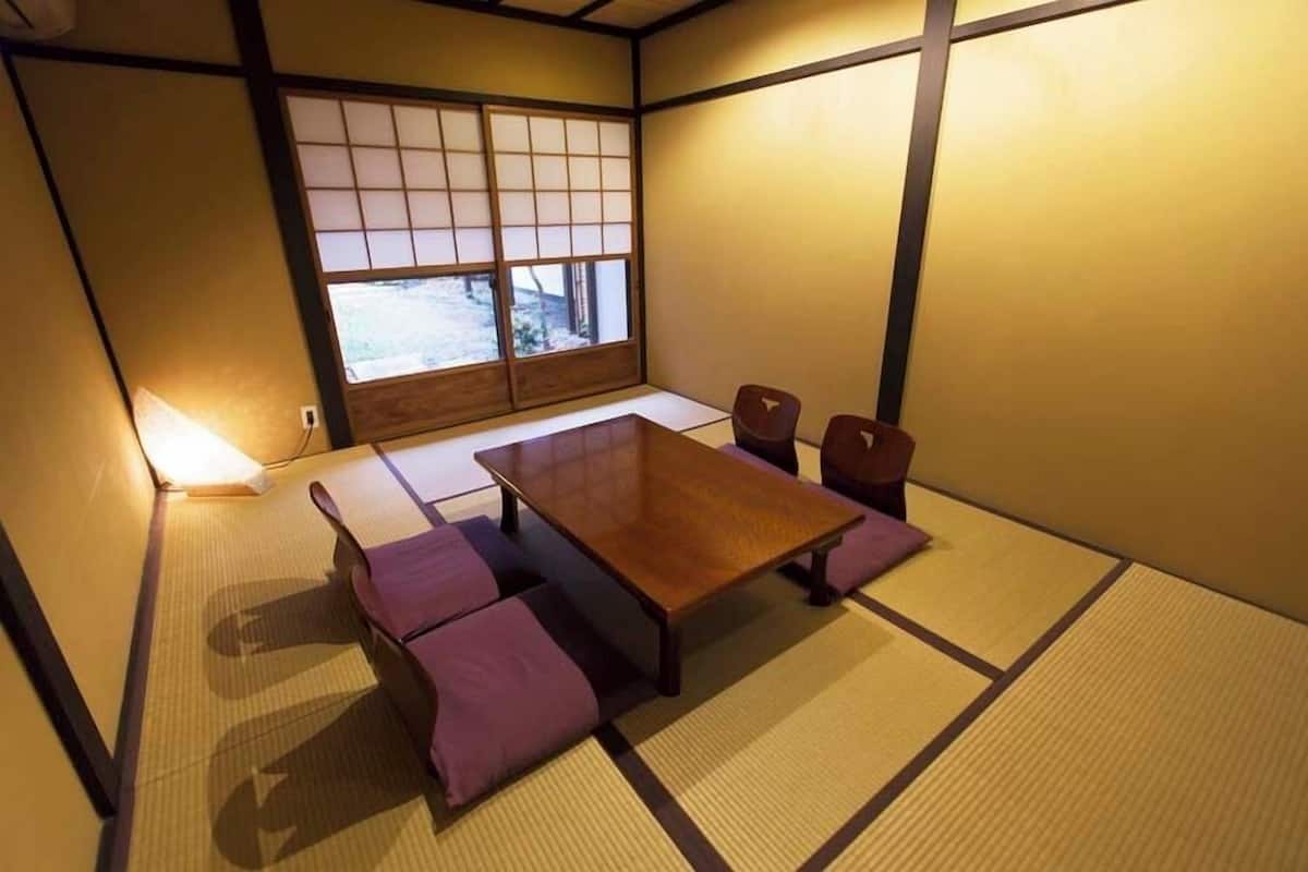 A tatami mat room with a wooden table and chairs, a window with a view, and a lamp.