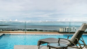 Une piscine séparée du reste par une paroi en verre, des chaises longues et une vue sur l’océan et une jetée.