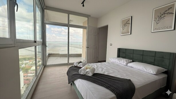Un dormitorio con un ventanal grande, una cama con ropa blanca, un cabecero verde y dos cuadros en la pared.