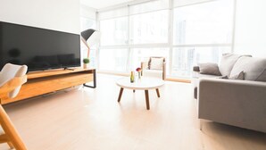 Ett modernt vardagsrum med en platt-TV, ett runt soffbord med blommor, en grå soffa och ett stort fönster med utsikt över staden.