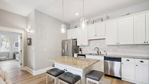 Eine moderne Küche mit weißen Schränken, Edelstahlgeräten und einer Arbeitsplatte aus Granit. Zwei Barhocker und Pendelleuchten befinden sich über der Kücheninsel.