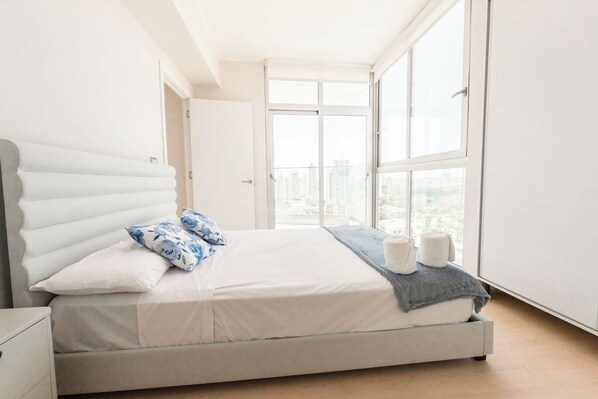 A modern bedroom with a large bed, a nightstand, and a view of the city through large windows.