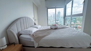 Ein ordentlich bezogenes Bett mit Kopfteil, ein Nachttisch mit Lampe und ein Fenster mit Blick auf die Stadt.