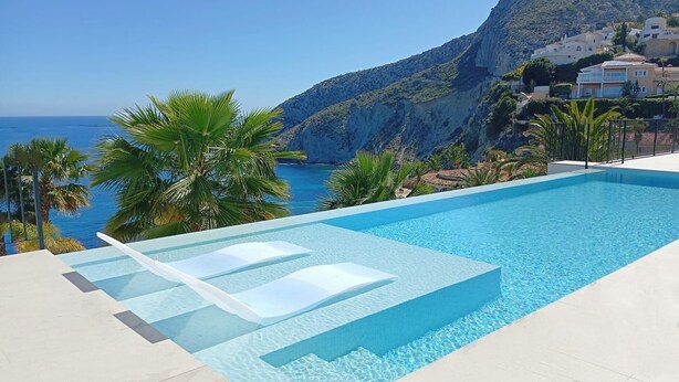 En swimmingpool med klart, blåt vand, omgivet af palmer og udsigt til hav og bjerge.