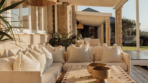 A covered outdoor seating area with a sofa and cushions, a table with a bowl, and a view of a building in the background. - Hinterland House (NSW)
