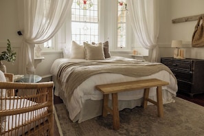 A bedroom with a bed, bench, wicker chair, and a dresser. - Hinterland House (NSW)