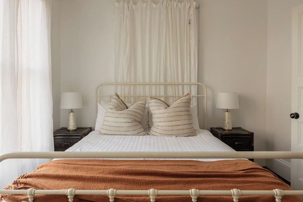 A bed with a metal frame, two bedside tables with lamps, a white curtain, and a brown blanket.