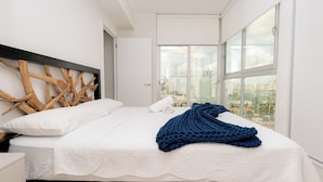 Een moderne hotelkamer met een groot bed, witte beddengoed, een houten hoofdbord en een stadszicht door grote ramen.