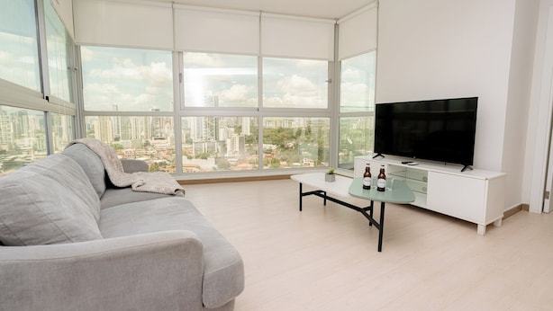 Een moderne woonkamer met een grijze bank, een glazen salontafel en een televisie op een witte kast. Grote ramen bieden uitzicht over de stad.