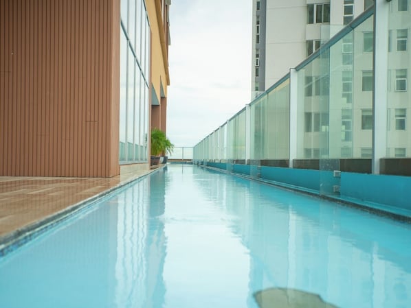 Una larga piscina interior con agua azul cristalina, rodeada de edificios modernos con grandes ventanales y un muro de madera en un lateral.