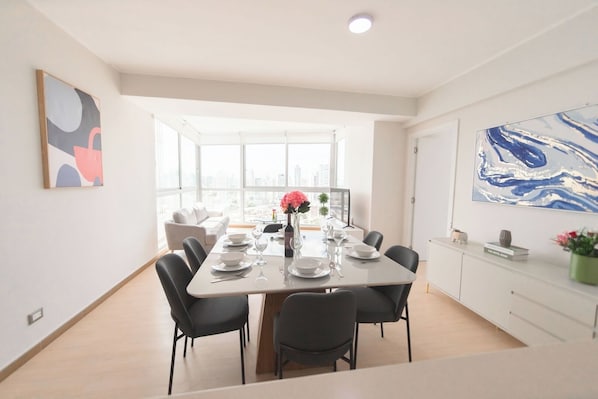 A modern dining area with a long table set for four, large windows offering a city view, and contemporary artwork on the walls.