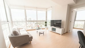A modern living room with a sofa, a coffee table, a TV on a white stand, and large windows offering a city view.