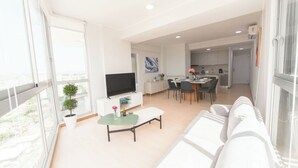 A modern apartment with a living room, dining area, and kitchen. There is a flat-screen TV on a white cabinet, a dining table with chairs, and a kitchen counter.
