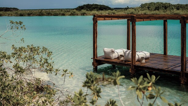 A wooden deck with white cushions and a small table, overlooking clear turquoise water. - Boca De Agua Bacalar (Felipe Carrillo Puerto)
