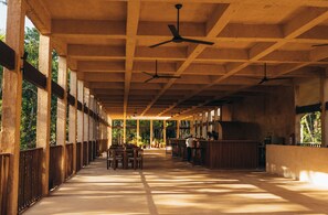Un espacio al aire libre cubierto con bancos y mesas de madera, ventiladores de techo y una zona de bar.