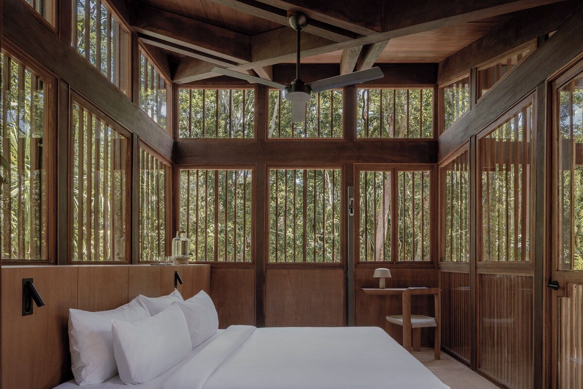 Una habitación de madera con una cama, un ventilador de techo y ventanas amplias.