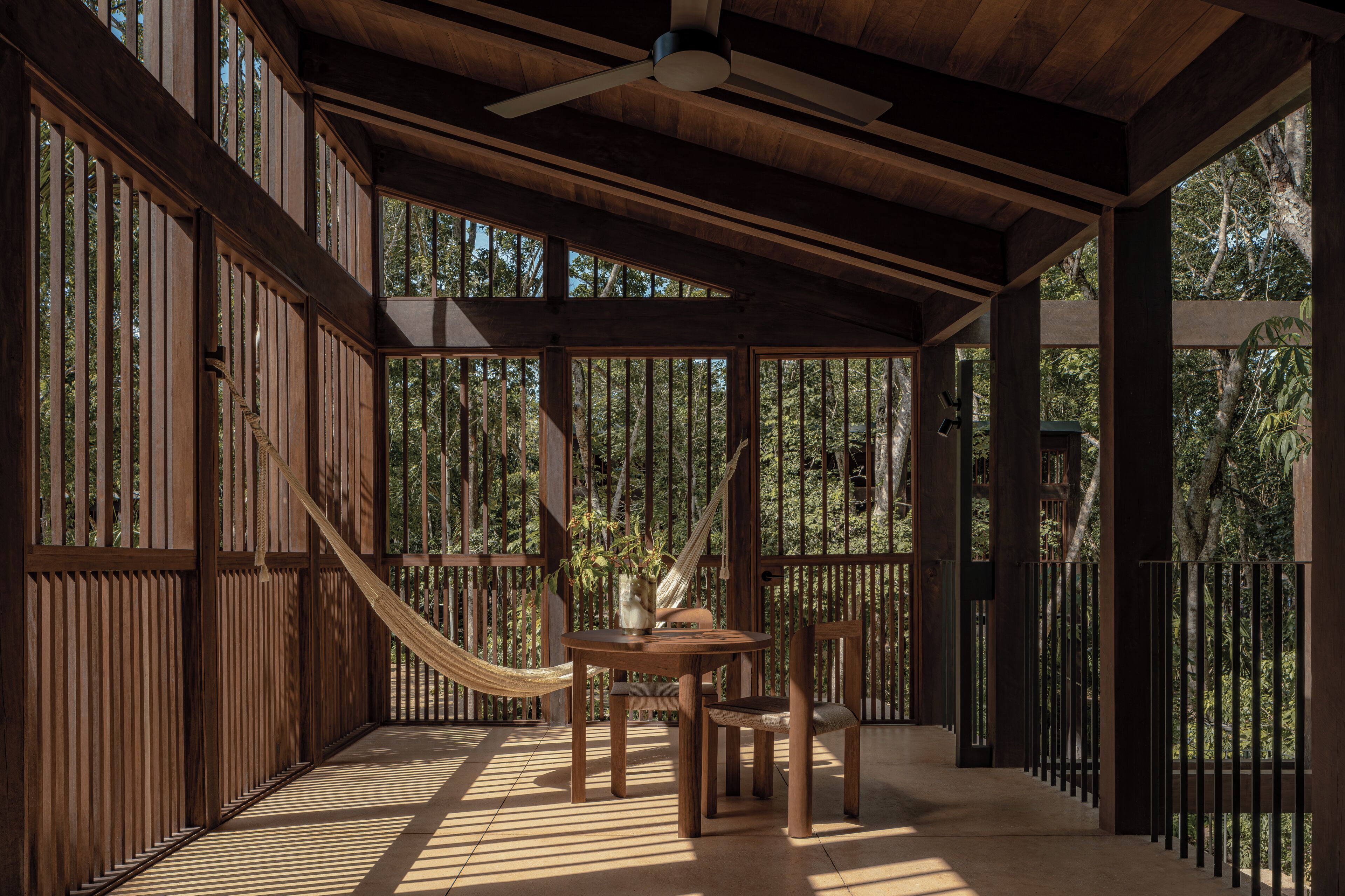 A wooden deck with a hammock, a table, and chairs, surrounded by vertical wooden slats and a view of trees.
