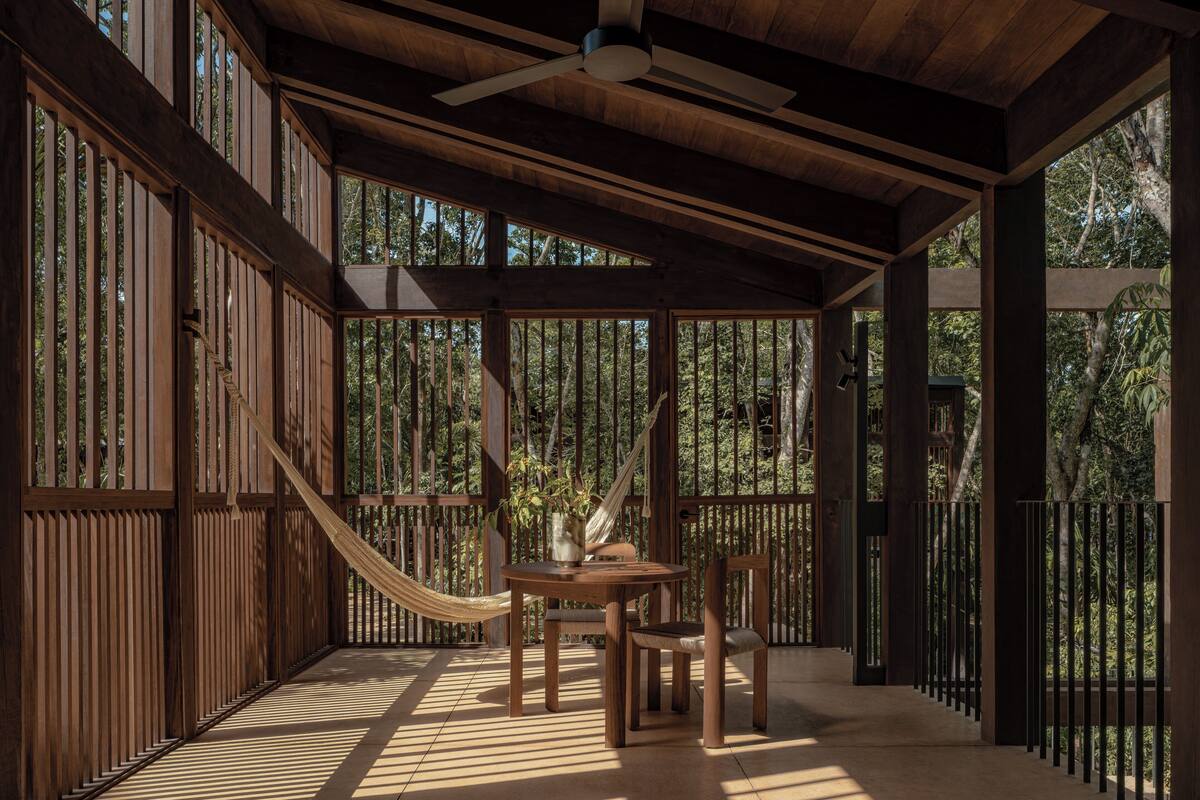 Una terraza de madera con una hamaca, una mesa y sillas, rodeada de listones verticales de madera y vistas a árboles.