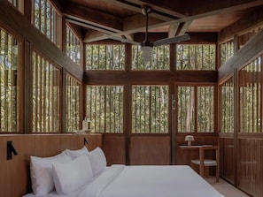 Una habitación de madera con una cama, un ventilador de techo y grandes ventanales con vista al bosque.
