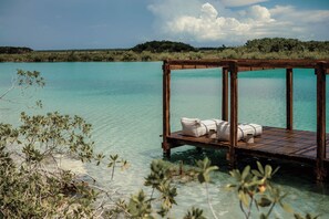 Une terrasse en bois agrémentée de coussins blancs et d’une petite table, offrant une vue sur une eau turquoise et cristalline.