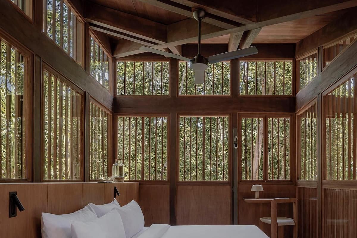 Una habitación de madera con una cama, un ventilador de techo y grandes ventanales con vista al bosque.