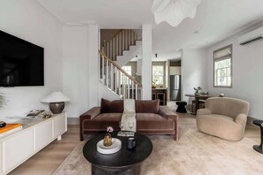 Ein modernes Wohnzimmer mit einem braunen Sofa, einem beigen Sessel und einem schwarzen Couchtisch. Im Hintergrund ist eine Treppe zu einem anderen Raum zu sehen.