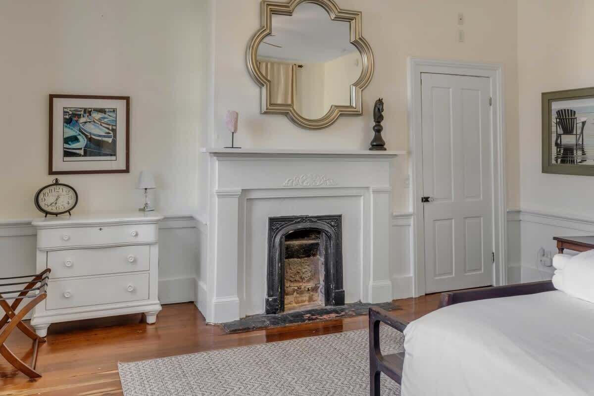 A bedroom with a fireplace, a bed, a dresser, a clock, and a framed picture on the wall.