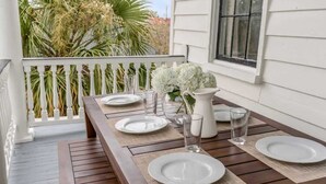 Mesa de comedor dispuesta con platos, vasos y una jarrón en una terraza de madera con barandilla blanca y vista a palmeras.