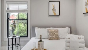 Een hotelkamer met een bed, een nachtkastje met een fles en glazen, een raam met zonwering en een ingelijst kunstwerk aan de muur.