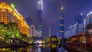 A nighttime cityscape with illuminated skyscrapers, a calm river reflecting the lights, and a traditional building with a red roof on the right. - Echerm Hotel Wuyangcun Metro Station (Guangzhou)