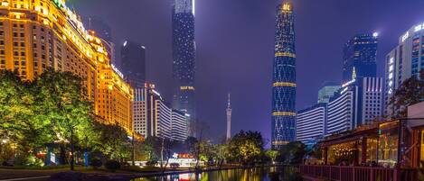 A nighttime cityscape with illuminated skyscrapers, a calm river reflecting the lights, and a traditional building with a red roof on the right.