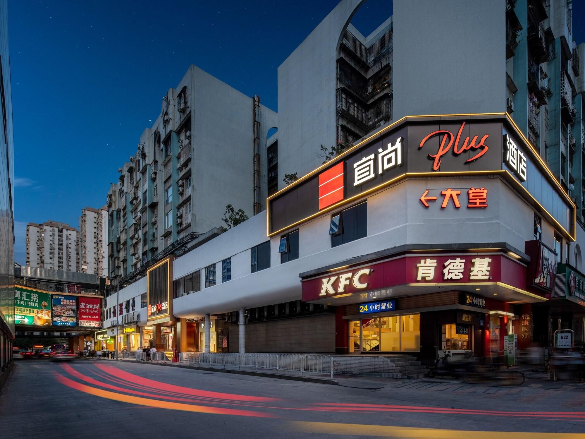 Un restaurante KFC con un letrero que indica servicio las 24 horas, ubicado en un edificio de varios pisos con una escalera visible y tiendas contiguas.