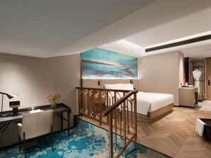Une chambre d’hôtel avec un lit, un bureau, une chaise, une petite table ornée d’un vase de fleurs et un tableau représentant un paysage marin accroché au mur.