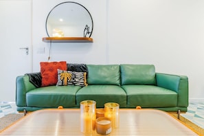 Een groene leren bank met gekleurde kussens, een ronde spiegel erboven en een tafel met kaarsen.