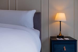 Un lit bien fait, avec du linge de lit blanc et une tête de lit sombre. À côté du lit, on trouve une petite table de chevet sur laquelle sont posés une lampe, un stylo et un bloc-notes.