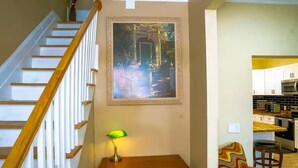 Una escalera con barandillas de madera, un cuadro enmarcado arriba, un escritorio con lámpara y una zona de cocina con armarios y electrodomésticos.