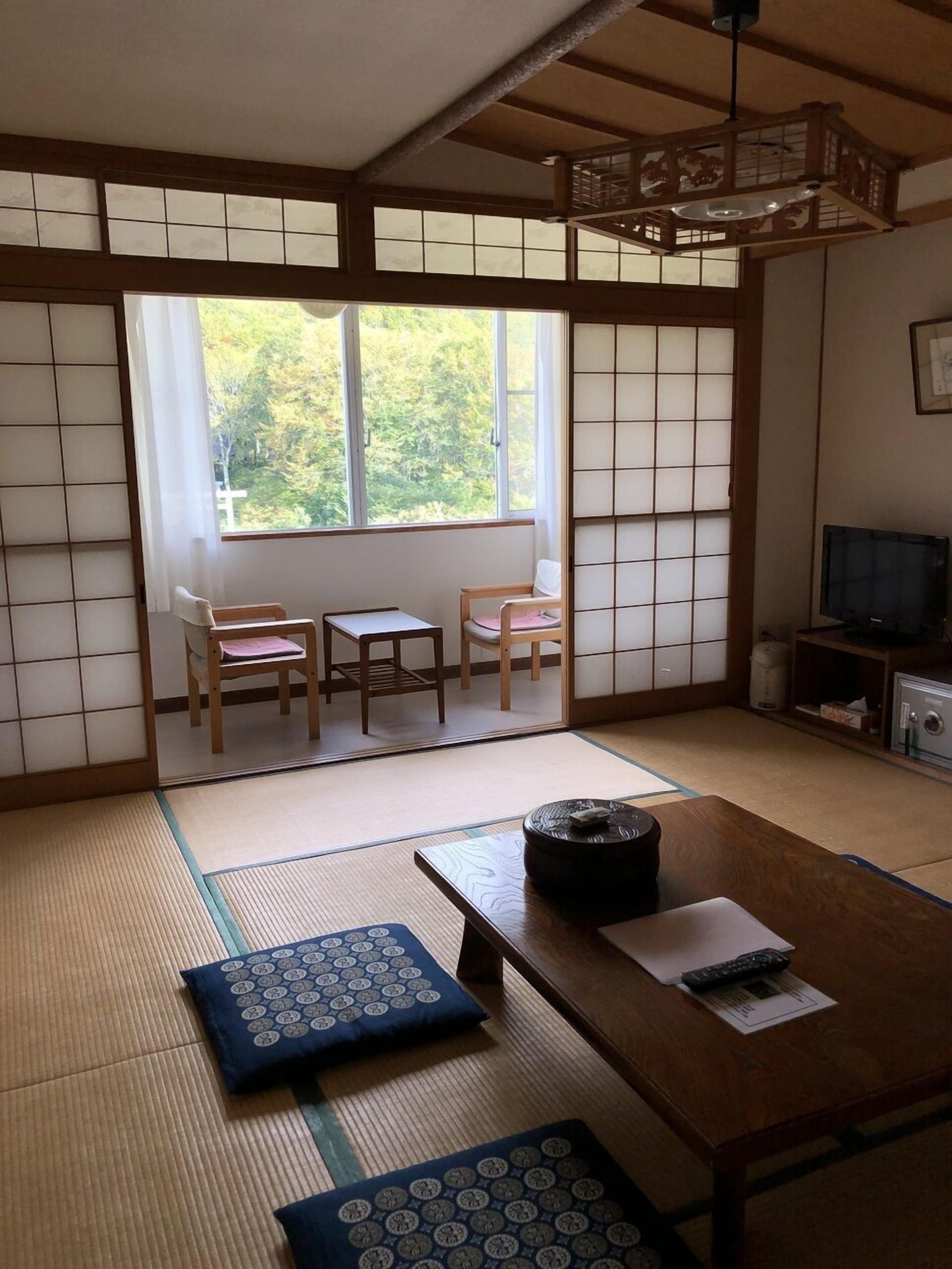 A traditional Japanese room with tatami flooring, a low table, two chairs, a television, and a sliding door leading to a balcony.
