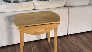 Un salon moderne avec un canapé d’angle, une table basse en bois, un ventilateur de plafond, un téléviseur à écran plat et une fenêtre équipée de stores.