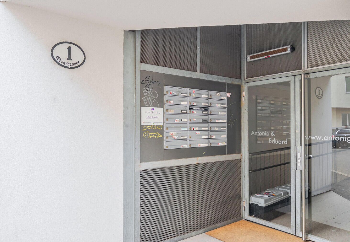 Ein Gebäudeeingang mit Glastür, Briefkasten und einer an der Wand befestigten Schilder.
