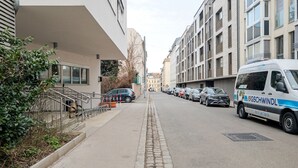 A street with parked cars and a van labeled 'Gschwindl'. Buildings line the street, and there is a sidewalk with a metal railing. - Blueground | Währing, view, nr Türkenschanzpark (Vienna)