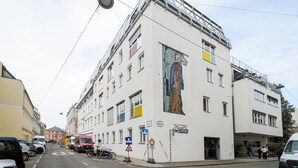 A multi-story building with a large mural on the facade, a ground-floor entrance, and a parking area with cars and bicycles. - Blueground | Währing, view, nr Türkenschanzpark (Vienna)