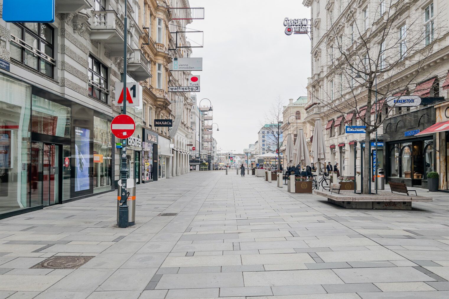 Uma rua pedonal larga, flanqueada por lojas e cafés, com uma loja MAX&Co. e uma placa Turek.