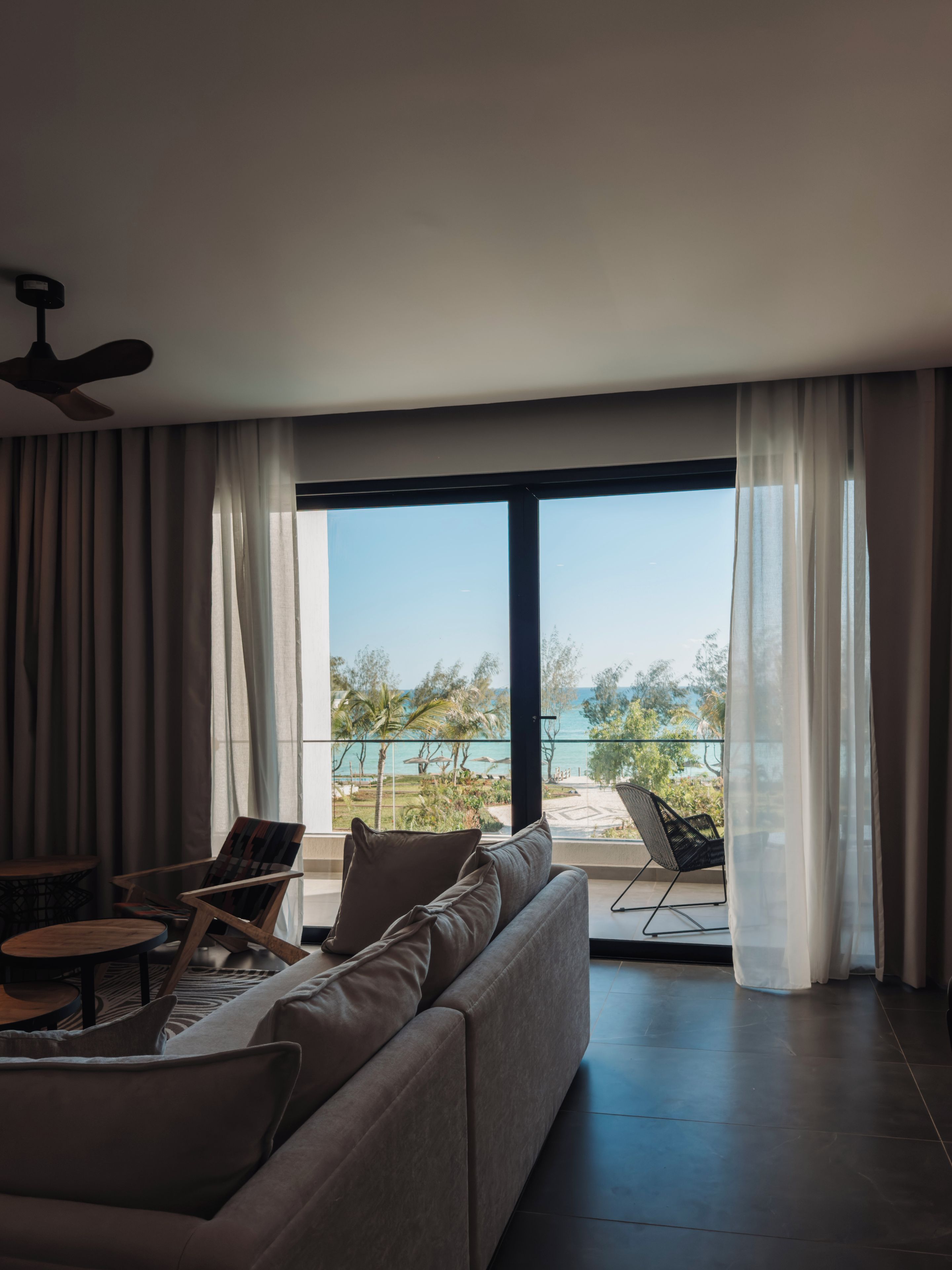 Uma sala de estar moderna com sofá, poltronas e uma mesa. É possível ver um ventilador de teto, e a sala possui janelas grandes com cortinas leves.