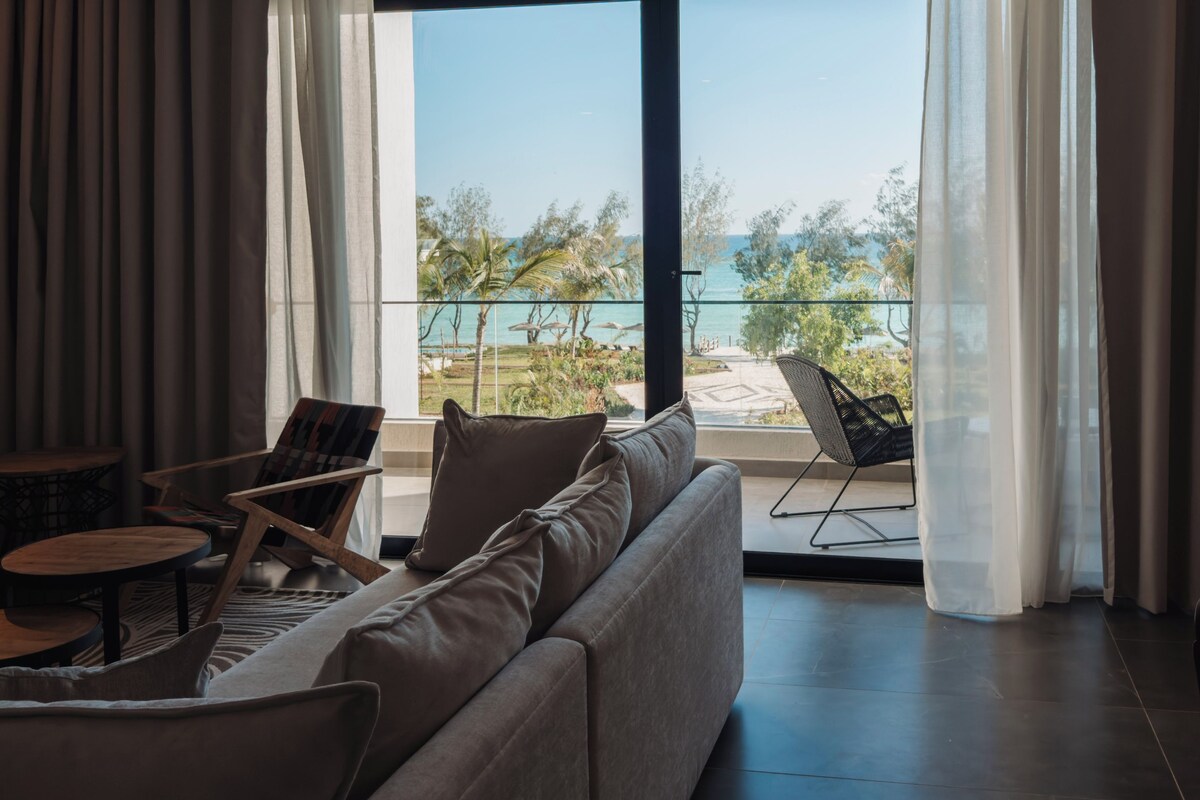 Una sala de estar moderna con un sofá, sillones y una mesa. Se ve un ventilador de techo, y la habitación tiene ventanas amplias con cortinas traslúcidas.